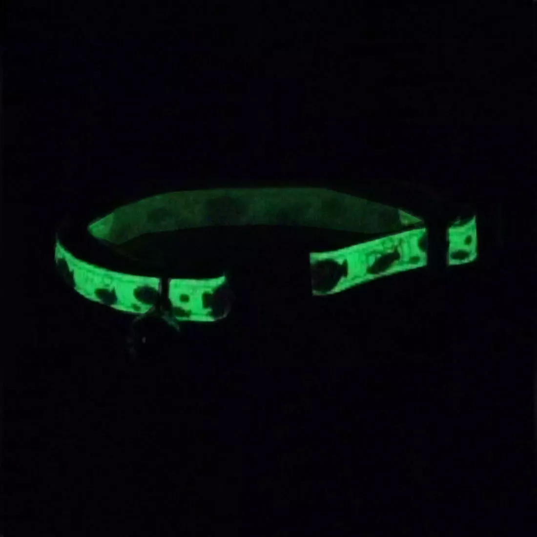 A product image of a glow‑in‑the‑dark adjustable breakaway collar displayed in low light. The collar strap glows bright green and shows repeating shapes, including fish and bubbles.