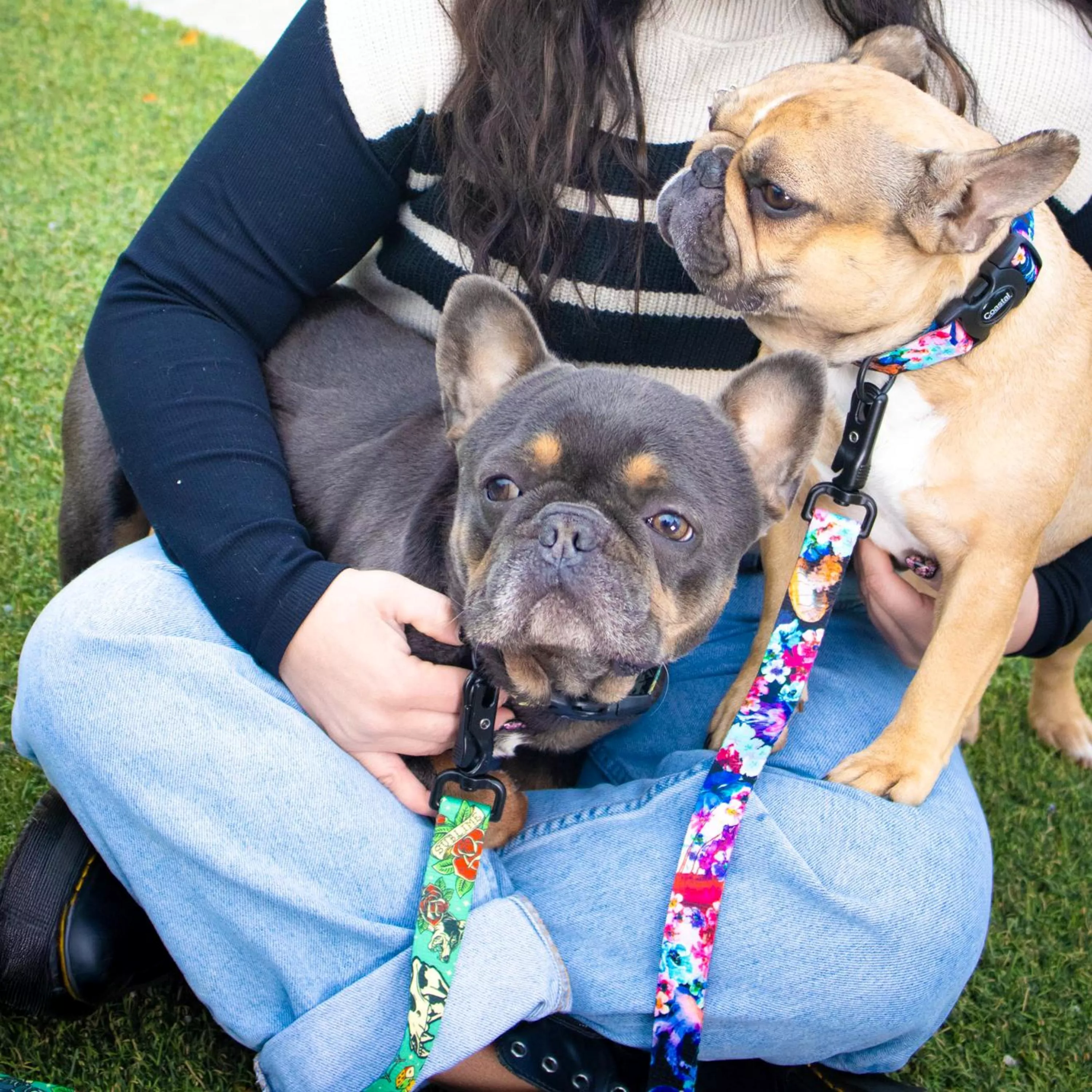Two dogs are sitting on a person’s lap outdoors on green grass. The person is wearing a black long-sleeve top with a cream-colored vest and light blue jeans. The dog on the left is wearing a Sublime adjustable collar and matching leash in the Tattoo Sleeve pattern, which features a teal background with colorful tattoo-inspired motifs such as red hearts, white skulls, and roses. The dog on the right is wearing a Sublime adjustable collar and matching leash in the Floral Jelly pattern, which displays a multicolor abstract floral design with shades of pink, purple, blue, teal, and orange on a dark background. Both leashes are attached to the collars with black metal clips.