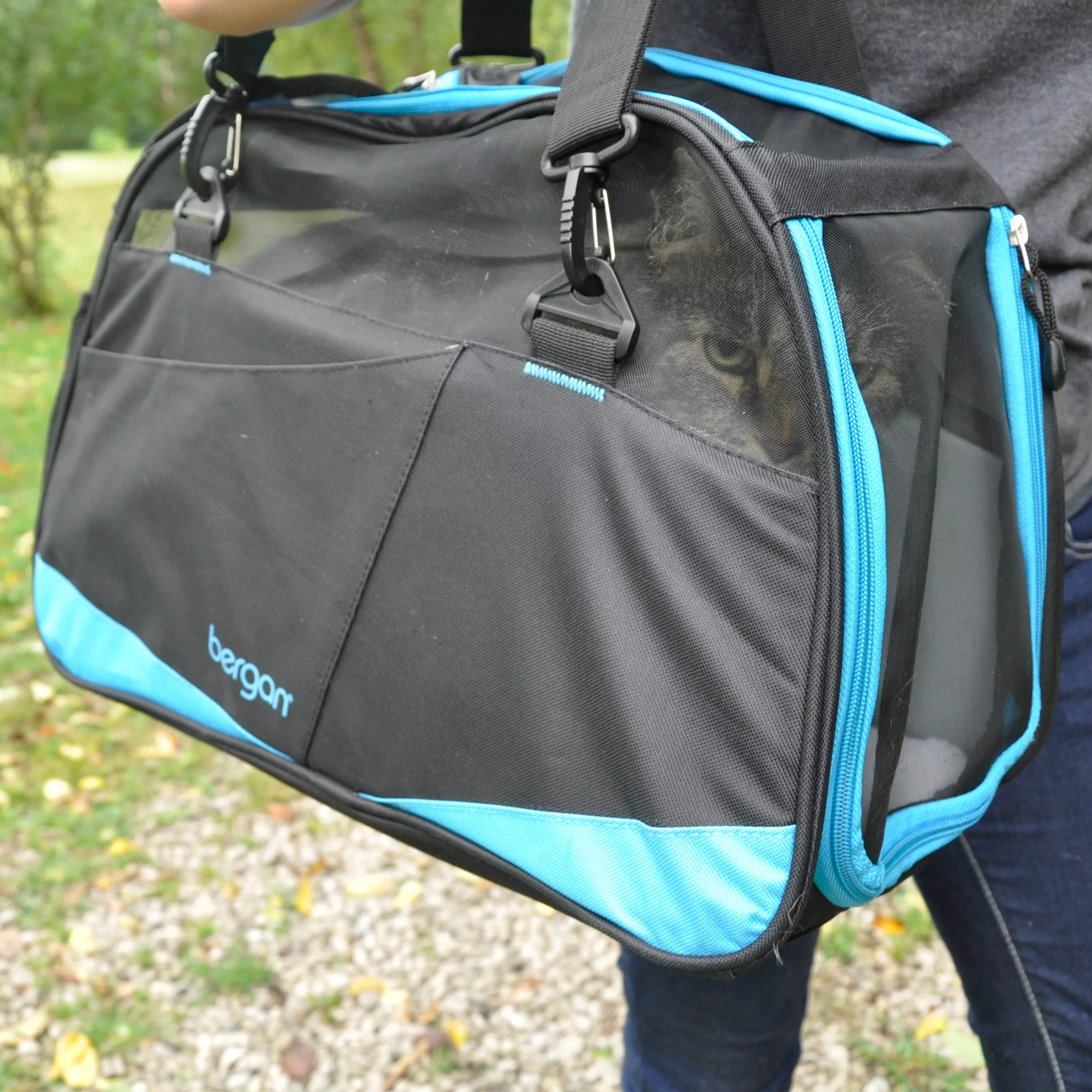 A person carries the Bergan® Comfort Carrier Voyager™ outdoors by its padded black handles. The carrier is primarily black with bright turquoise trim that outlines the edges, zipper seams, and front accessory pocket. The side panel features a large curved mesh ventilation window in black, framed by turquoise piping. The front of the carrier includes a wide slip pocket with a turquoise accent along the bottom edge. Reinforced stitching and structured fabric panels give the carrier a stable shape. The background includes grass, gravel, and natural outdoor lighting.