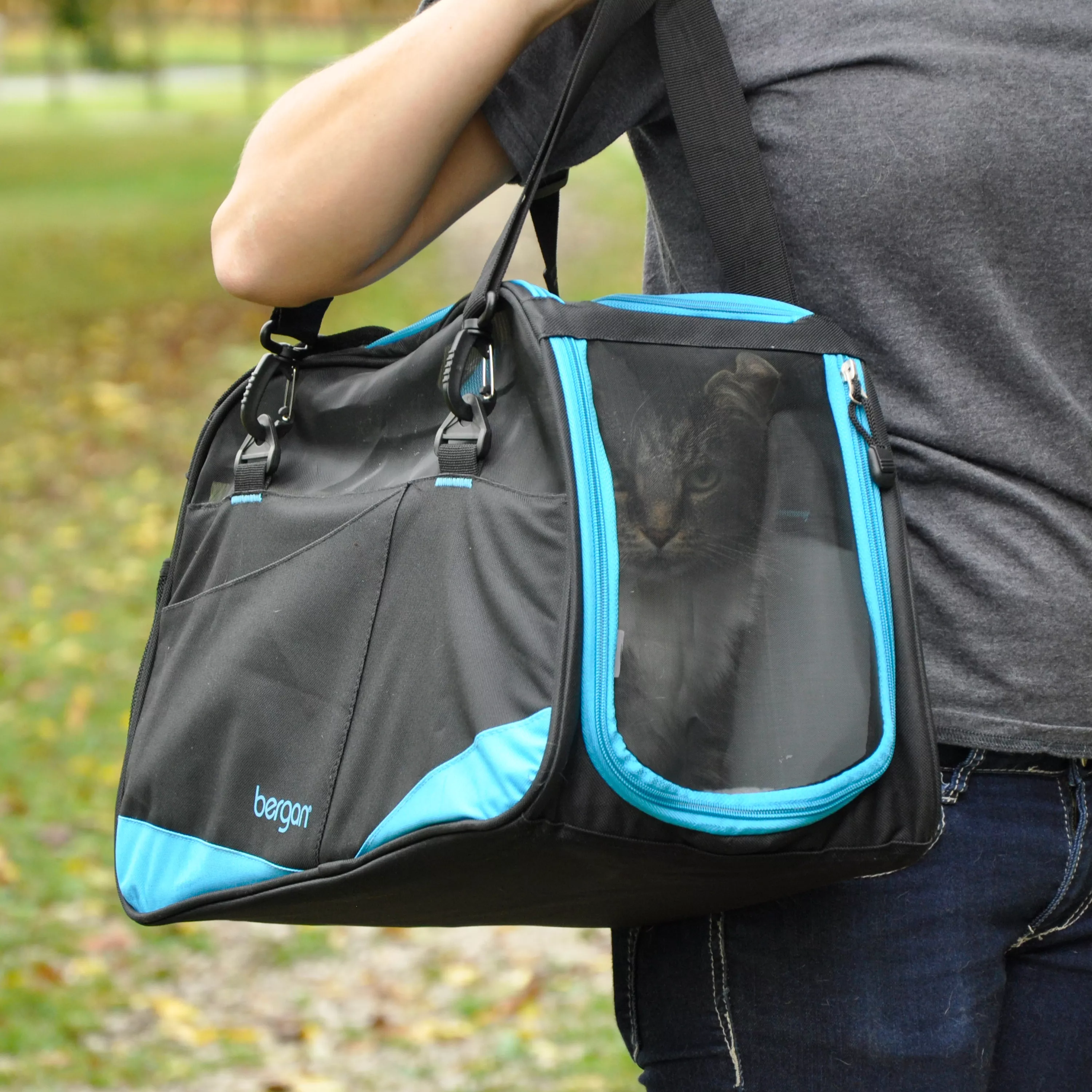 A person carries the Bergan® Comfort Carrier Voyager™ outdoors by its padded black handles. The carrier is primarily black with bright turquoise trim that outlines the edges, zipper seams, and front accessory pocket. The side panel features a large curved mesh ventilation window in black, framed by turquoise piping. The front of the carrier includes a wide slip pocket with a turquoise accent along the bottom edge. Reinforced stitching and structured fabric panels give the carrier a stable shape. The background includes grass, gravel, and natural outdoor lighting.