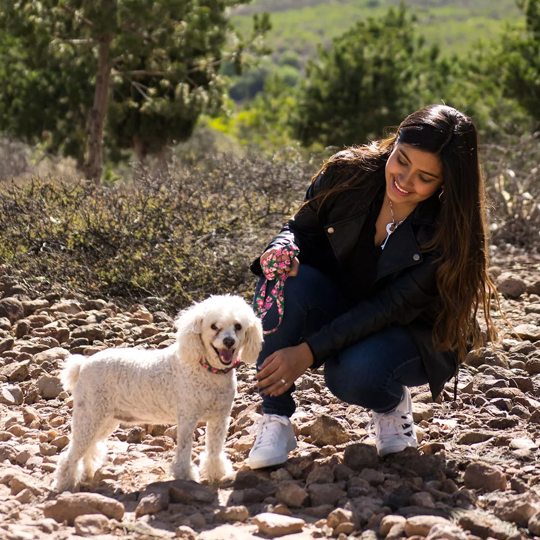 A lifestyle image of a dog wearing an adjustable collar and leash set with a tropical flower pattern. The collar strap has a green background with pink and white tropical flowers. The buckle is red plastic, and a silver metal D‑ring attaches to the matching leash. The dog has light curly fur and is sitting outdoors next to a person on a grassy hillside.